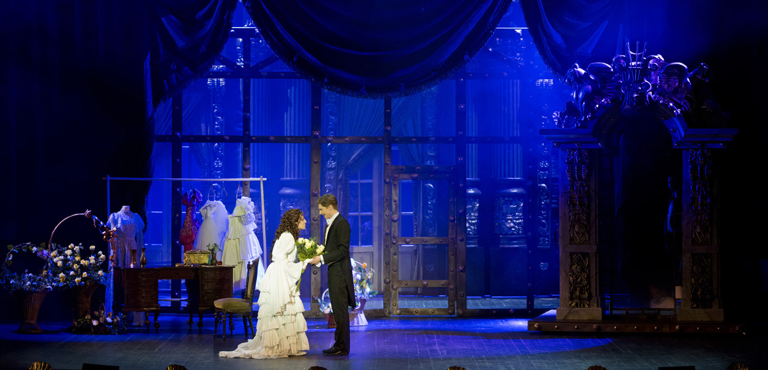 Scena oświetlona niebieskim światłe. Na środku kobieta i mężczyzna stoją zwróceni do siebie, trzymją sie za ręce.Po prawej stronie stoi duże lustro, po lewej stojak z sukienkami i kosze z kwiatami.