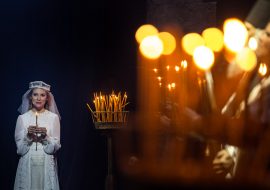 Na zdjęciu kobieta w białej, koronkowej sukience i w welonie. Trzyma w dłoniach zapaloną świeczkę. Na środku, na stojaku stoi kilkadziesiąt świeczek, część z nich pali się. Po prawej stronie w rozmytym tle, widać kilka osób trzymających zapalone świeczki.