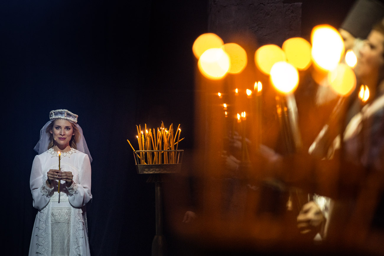 Na zdjęciu kobieta w białej, koronkowej sukience i w welonie. Trzyma w dłoniach zapaloną świeczkę. Na środku, na stojaku stoi kilkadziesiąt świeczek, część z nich pali się. Po prawej stronie w rozmytym tle, widać kilka osób trzymających zapalone świeczki.