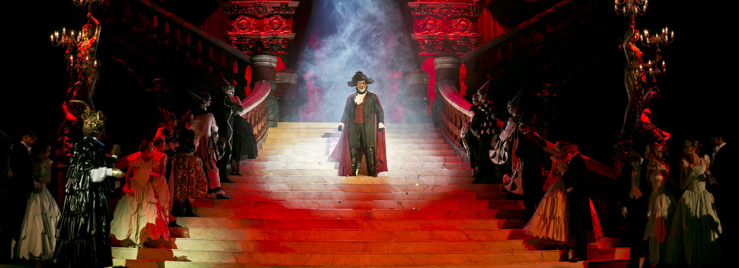 Scena oświetlona czerwonym światłem. Na środku dużych schodów stoi mężczyzna w czerwonym płaszczu i kapeluszu, z maską na twarzy. Po obydwu stronach, na stopniach schodów stoją kobiety i mężczyźni w balowych strojach.