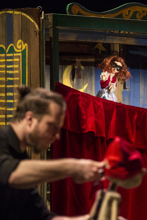 Na przodzie mężczyzna w czarnej koszuli, trzyma drewnianą lalkę. Dalej teatrzyk kukiełkowy. Na czerwonej zasłonie lalka teatralna.