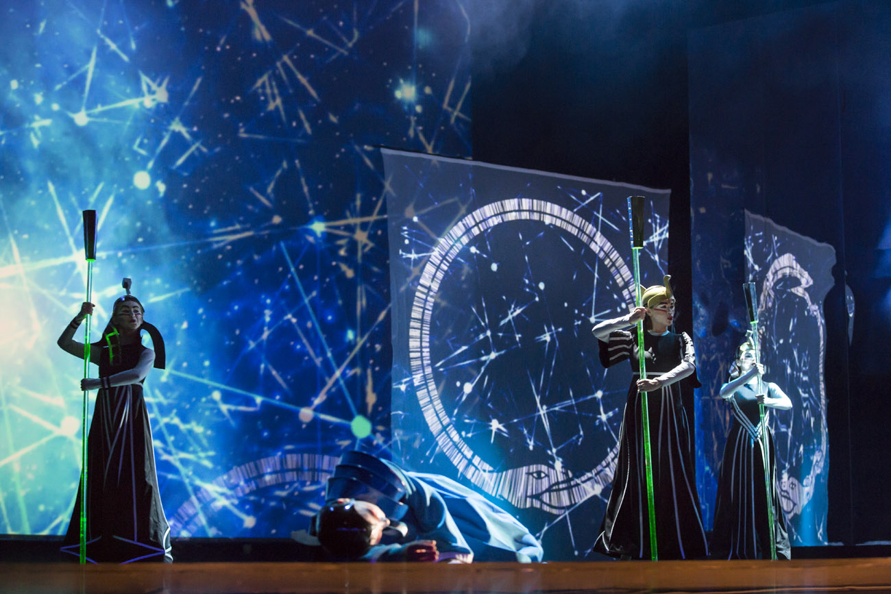 Na scenie stoją trzy postacie w czarnych, długich kostiumach. Trzymają w rękach długie podświetlone na zielono  Przed nimi lezy mężczyzna w niebieskim ubraniu.