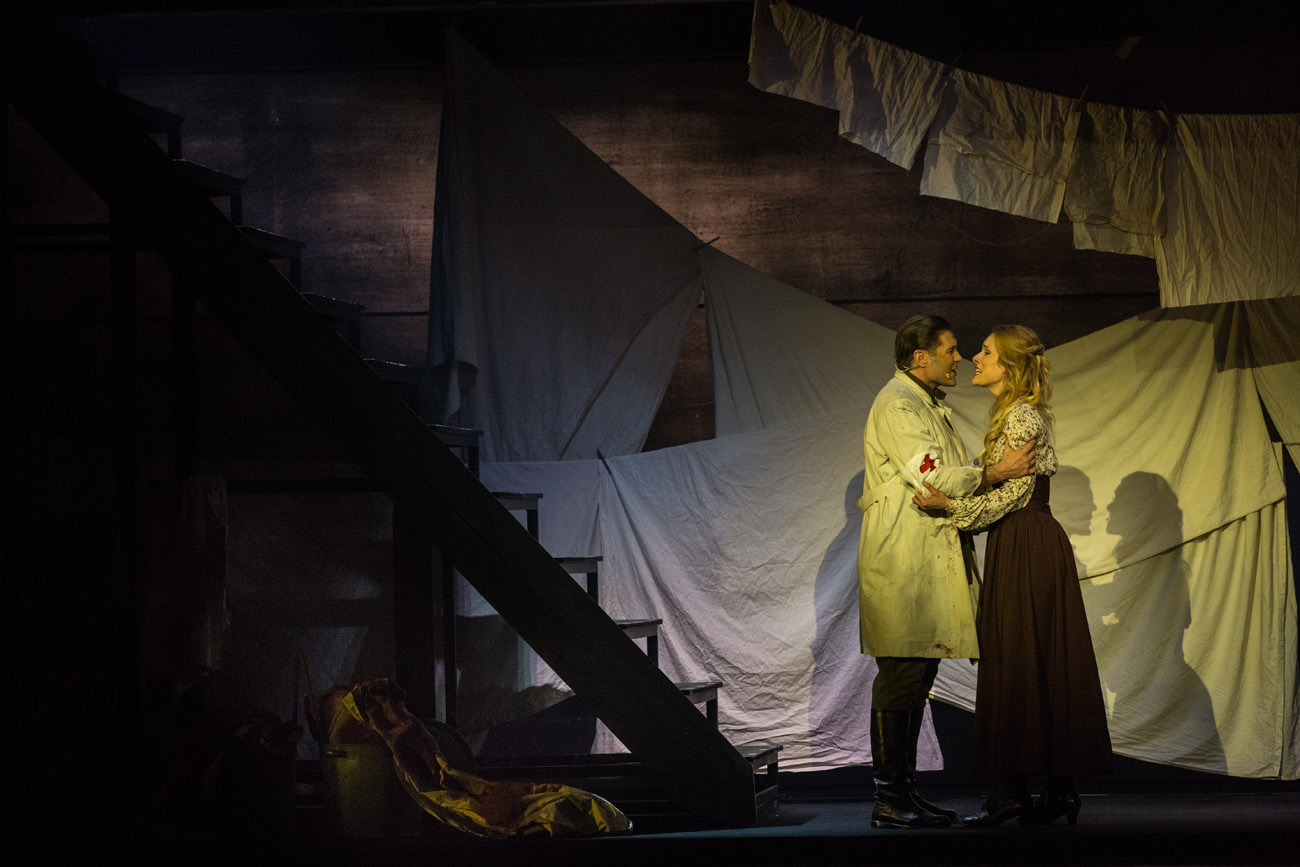 Na środku kobieta i mężczyzna w białym fartuchu, z opaską na ramieniu. Na opasce czerwony krzyż. Stoją przodem do siebie, trzymając się za ramiona. Za nimi , na różnych wysokościach, wiszą rozłożone białe prześcieradła. Po lewej stronie schody.