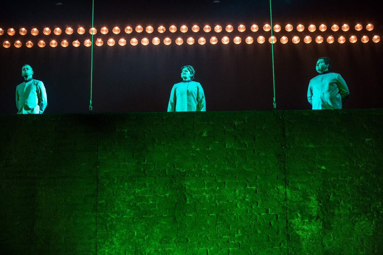 Na zdjęciu mur. Z góry zwisają dwie liny. Pomiędzy nimi stoi trzech mężczyzn. Na górze świecą dwa rzędy lamp.