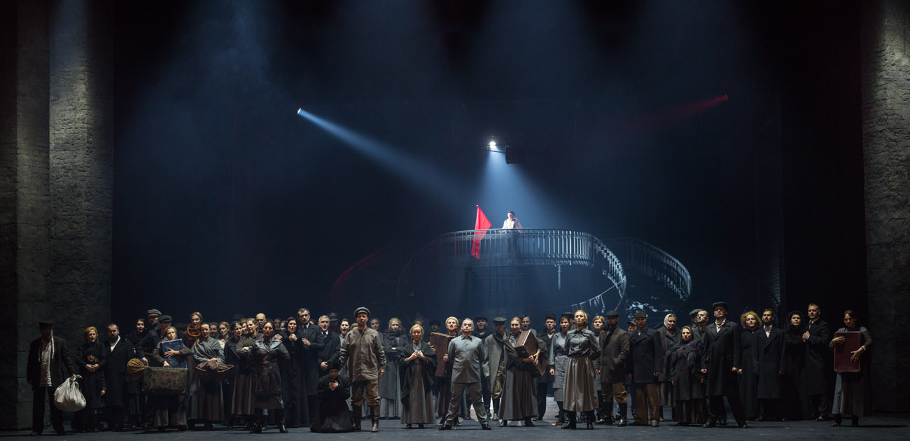 Na scenie kilkadziesiąt osób.Kobiety i mężczyźni. Stoją w rzędzie. Kilkoro z nich trzyma walizki. Jeden z nich klęczy. Za nimi wysokie schody, na nich stoi mężczyzna z flagą w kolorze czerwonym.