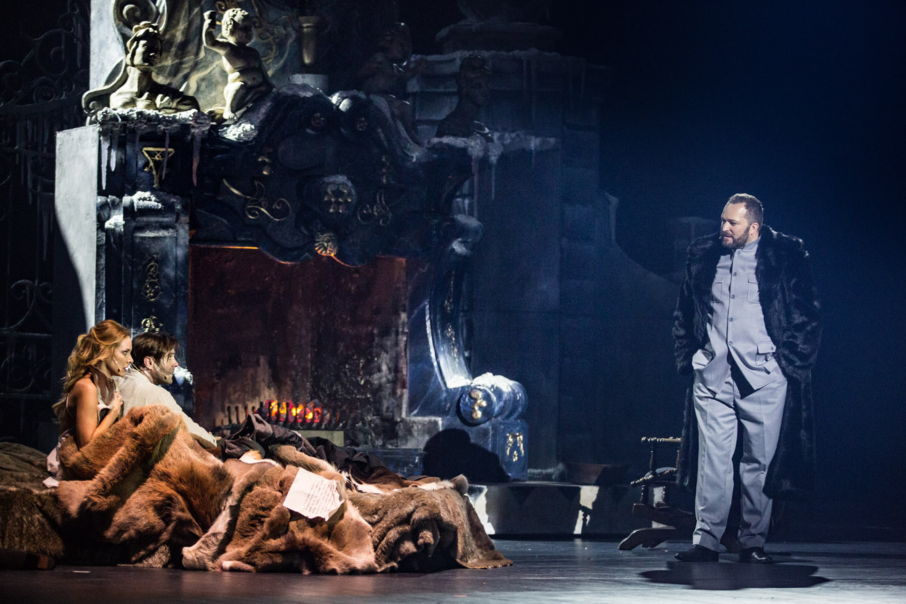 Po lewej stronie sceny kobieta i mężczyzna przykryci dużym futrem. Za nimi tlący się kominek. Po prawej mężczyzna w czarnym, futrzanym płaszczu, patrzy w kierunku pary.