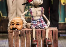 Na środku, na drewnianej skrzynce siedzi drewniana lalka w spodenkach w kratkę i czerwonej czapce. Obok leży maska. W tle dwie osoby trzymające róże.