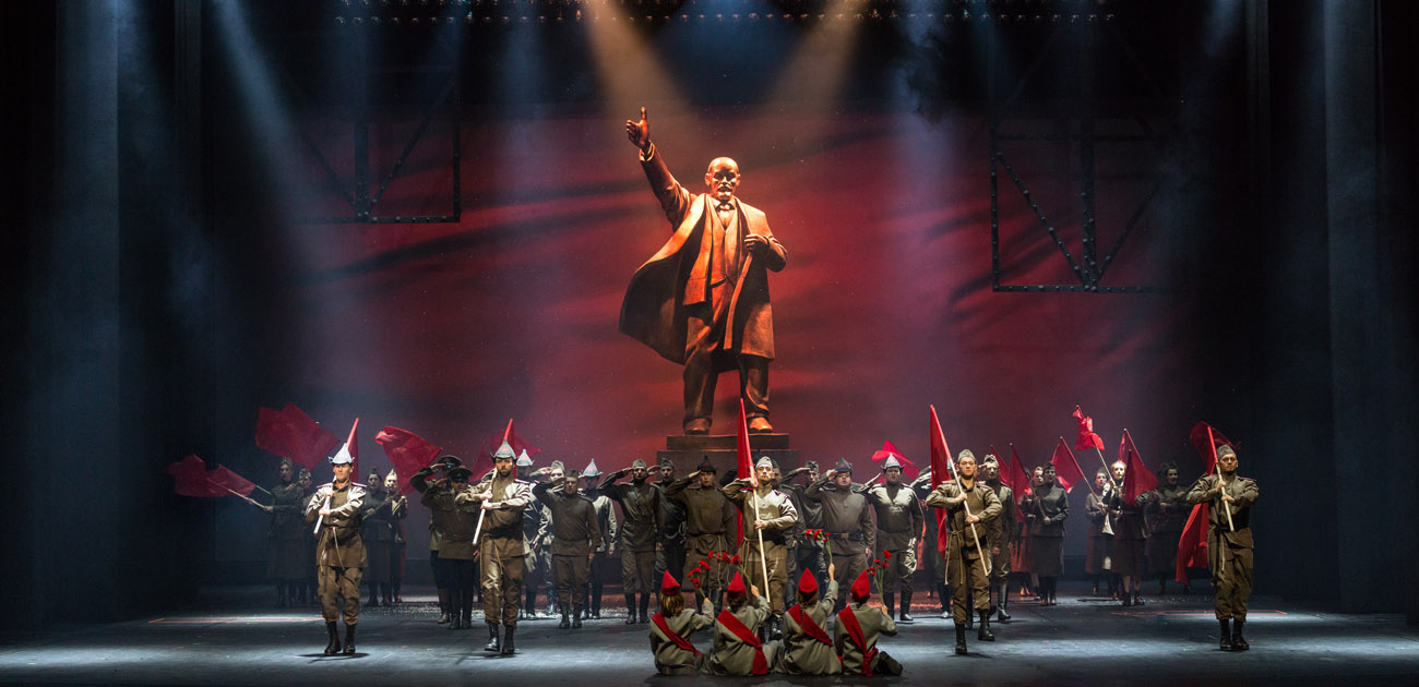 Na scenie mężczyźni i kobiety w mundurach. Tyłem do widowni siedzą cztery kobiety, w rękach trzymają pdniesione do góry kwiaty. Dalej , w rzędzie kilku mężczyzn trzyma oparte na ramionach flagi w kolorze czerwonym. Za nimi grupa mężczyzn, wszyscy stoją na baczność  trzymając dłonie przy skroni w geście salutowania. Wśród nich kilka kobiet z flagami w kolorze czerwonym. Na końcu, na środku, pomnik Lenina trzymającego dłoń w górze.