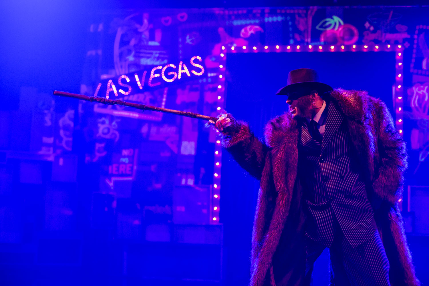 Scena oświetlona niebieskim światłem. Na środku stoi mężczyzna w garniturze i futrze, w ręku trzyma laskę. W oddali widać podświetlony napis " Las Vegas"