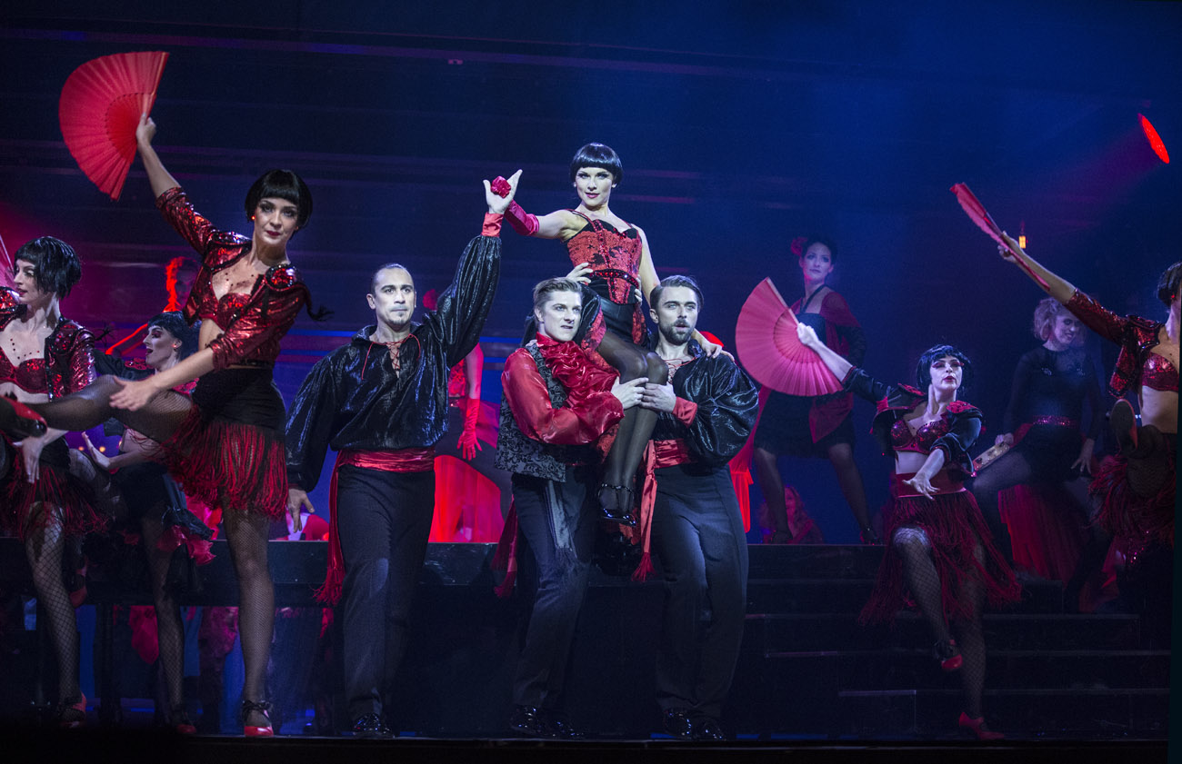 Grupa kobiet i mężczyzn w czerwono-czarnych hiszpańskich kostiumach tańczy na scenie. Na środku dwóch mężczyzn trzyma na ramionach kobietę. Po obydwu stronach, kobiety z rozłożonymi, czerwonymi wachlarzami.