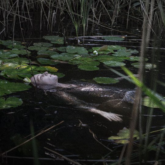 Na zdjęciu kobieta zanurzona w wodzie, twarzą do góry. Dookoła liście kwiatów wodnych.