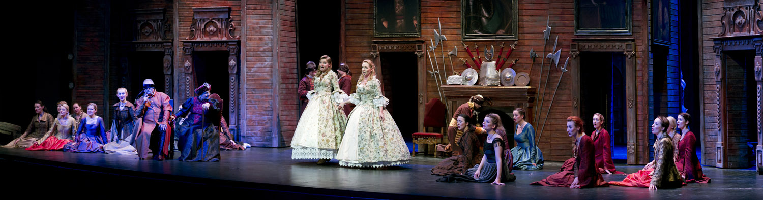 Dwie kobiety w długich , balowych sukniach stoją na środku sceny. Śpiewają. Po prawej i polewej stronie siedzą kobiety i mężczyźni