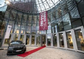 Wejście do Opery i Filharmonii Podlaskiej. Przed wejściem czerwony dywan.Po lewej stronie samochód marki BMW. Na górze zwisający w dół billboard z napisem: ''Baron cygański. Johann Strauss''.
