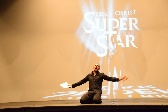 Krzysztof Gorczak bierze udział w castingu do spektaklu „Jesus Christ Superstar”. Ubrany w ciemne kolory klęczy na ziemi, ma rozłożone ręce, w prawej trzyma partyturę, śpiewa fragment piosenki ze spektaklu. W tle widoczny jest biały logotyp spektaklu „Jesus Christ Superstar” na pomarańczowym tle.